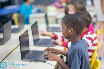 A child working on a laptop in a classroom with other children doing the same. Data Protection Education logo in the bottom right of the picture